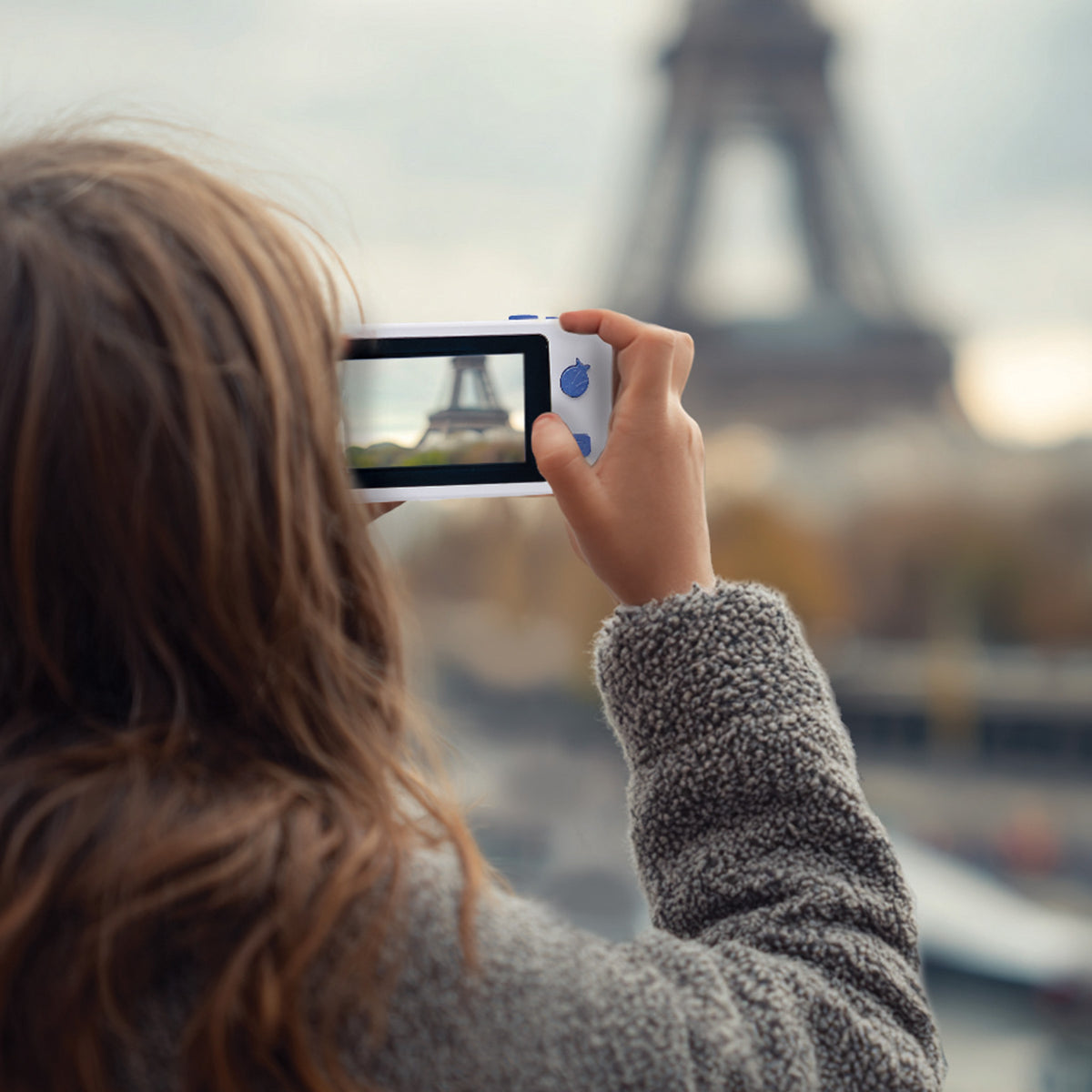 A woman with long brown hair takes a photo of the Eiffel Tower in Paris using a white handheld digital camera with blue buttons, seen from behind.