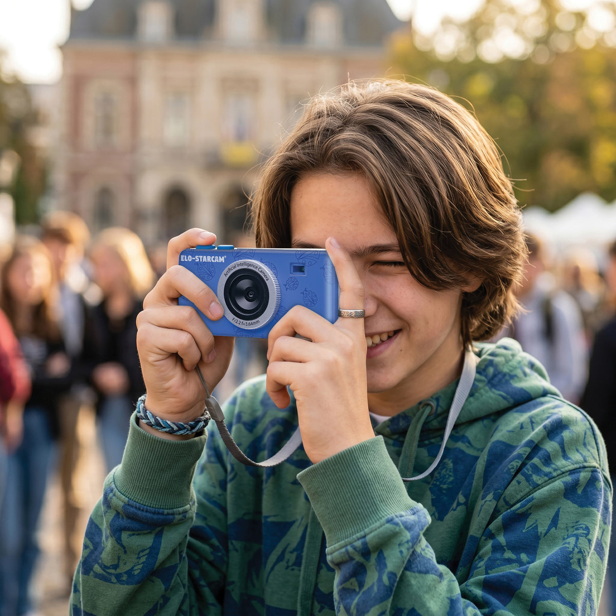 A smiling young person outdoors holding a blue Lexibook Starcam AI camera up to their eye, wearing a green and blue patterned hoodie with a blurred crowd and building in the background.